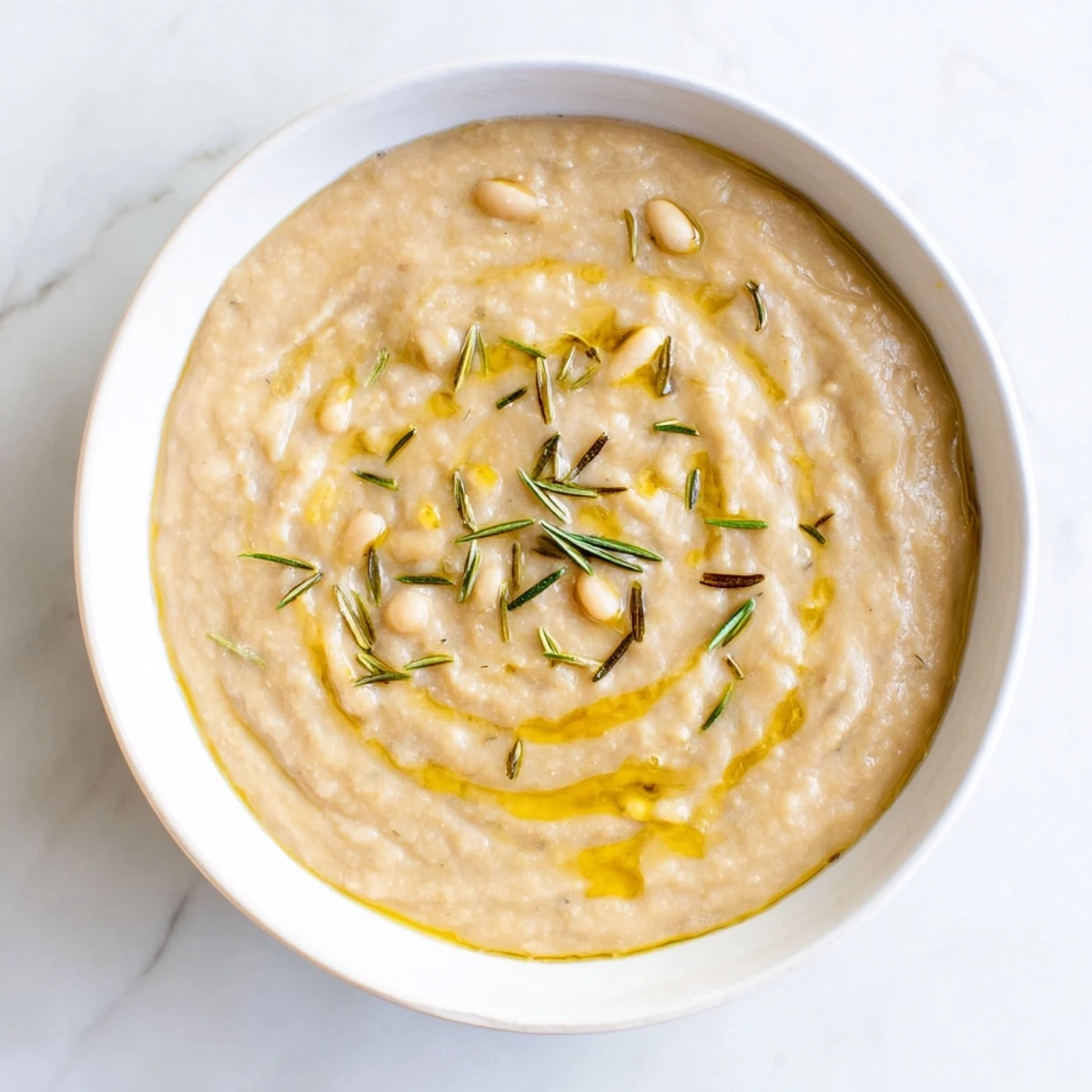 Enjoy a steaming bowl of Creamy White Bean and Rosemary Soup, garnished with fresh rosemary and a drizzle of olive oil.