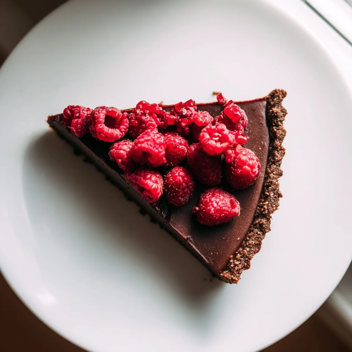 Overhead view of a slice of Decadent dark chocolate tart with raspberries, featuring powdered sugar dusting and a gooey center on a white plate.
