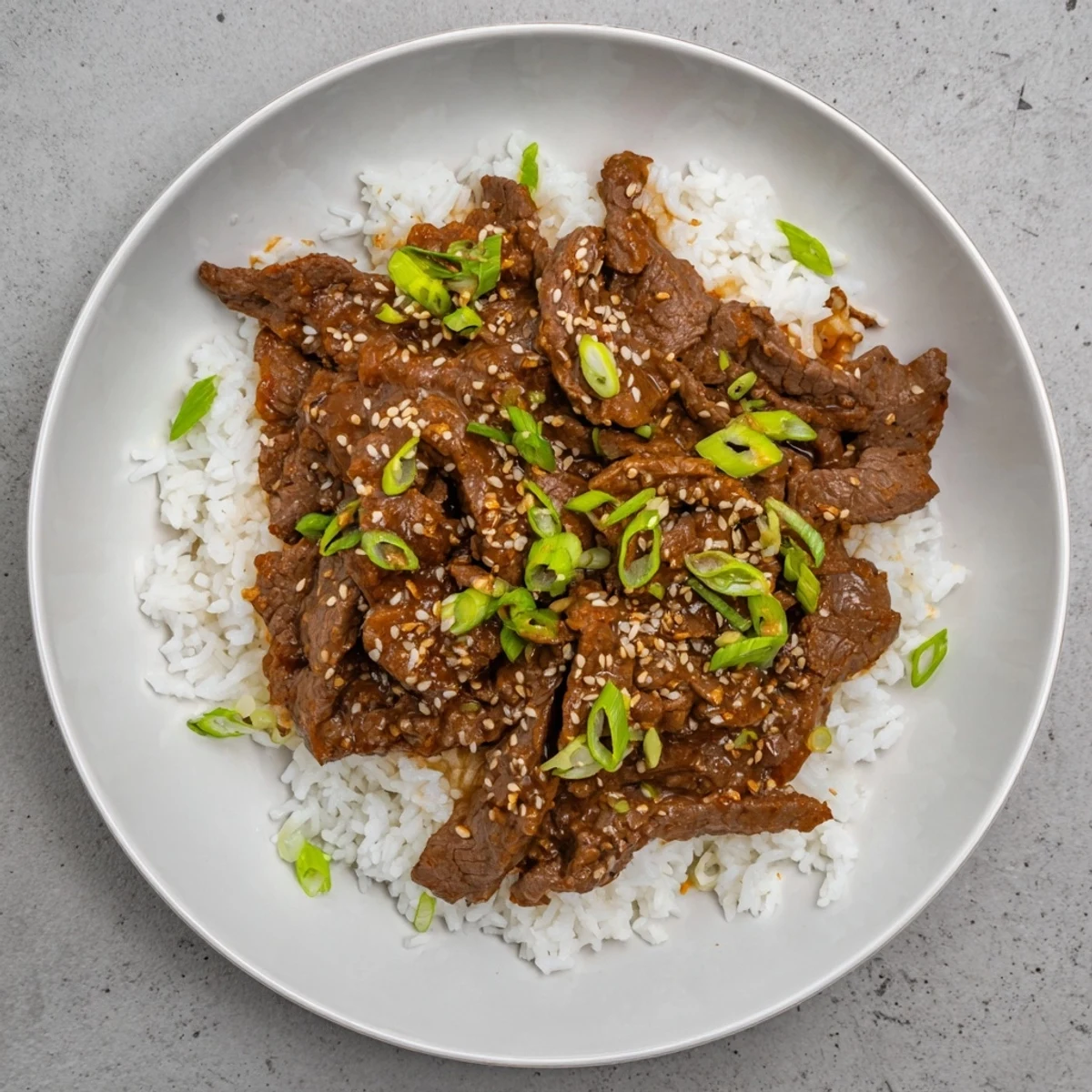 Steamed jasmine rice topped with glossy Ultimate Slow Cooker Mongolian Beef, garnished with green onions and sesame seeds.