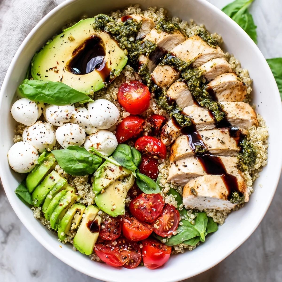 Juicy pesto-marinated chicken slices rest atop fluffy quinoa with fresh mozzarella, cherry tomatoes, and avocado in a grain bowl.