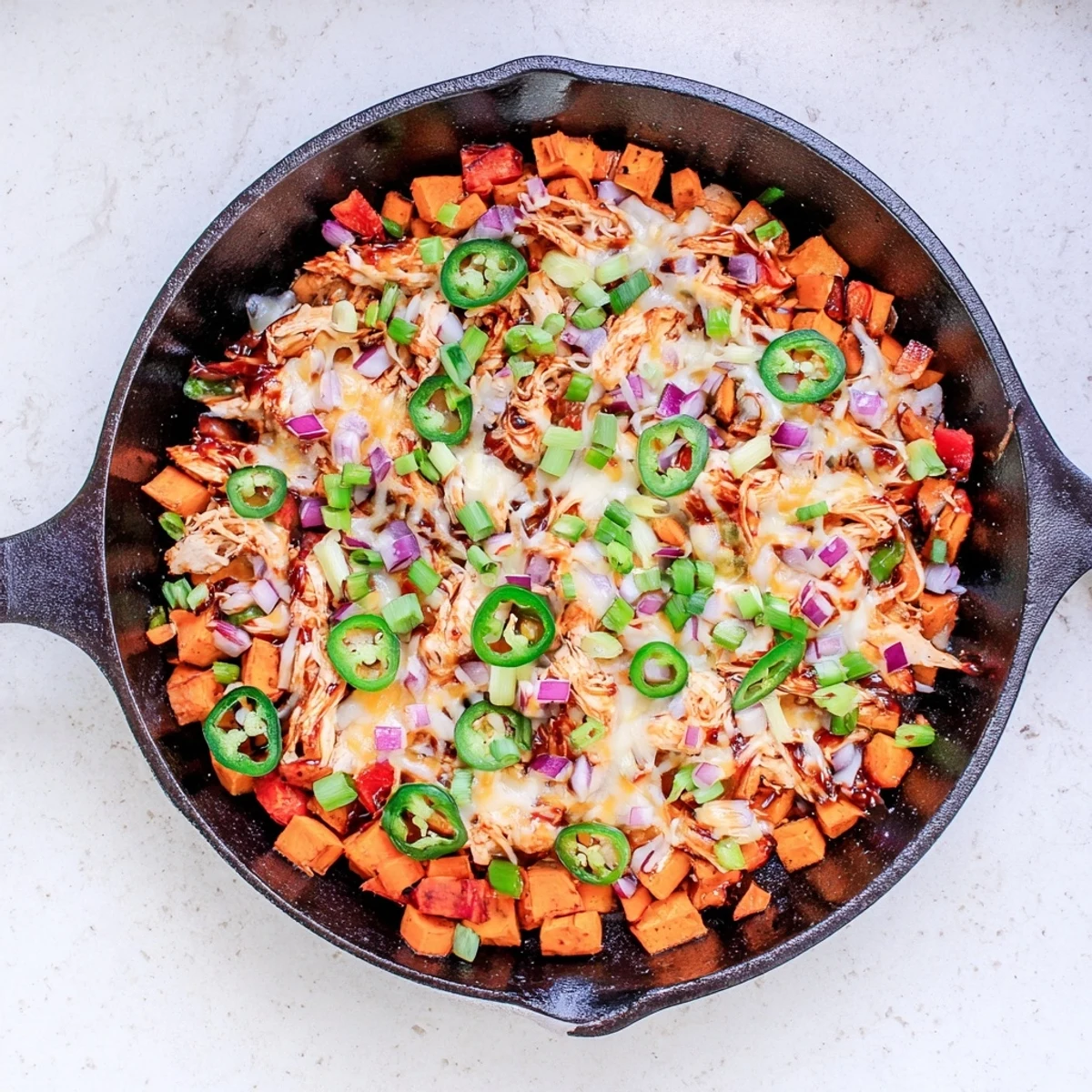 Golden melted cheese bubbling over smoky BBQ chicken and sweet potatoes in a sizzling cast iron skillet, garnished with green onions and jalapeños. Sweet Potato Skillet with BBQ Chicken & Cheese served hot.